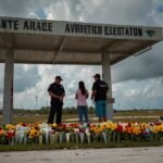A somber gathering at a convenience store on the Miccosukee Indian Reservation, with community members placing flowers and candles at a makeshift memorial under a cloudy sky.